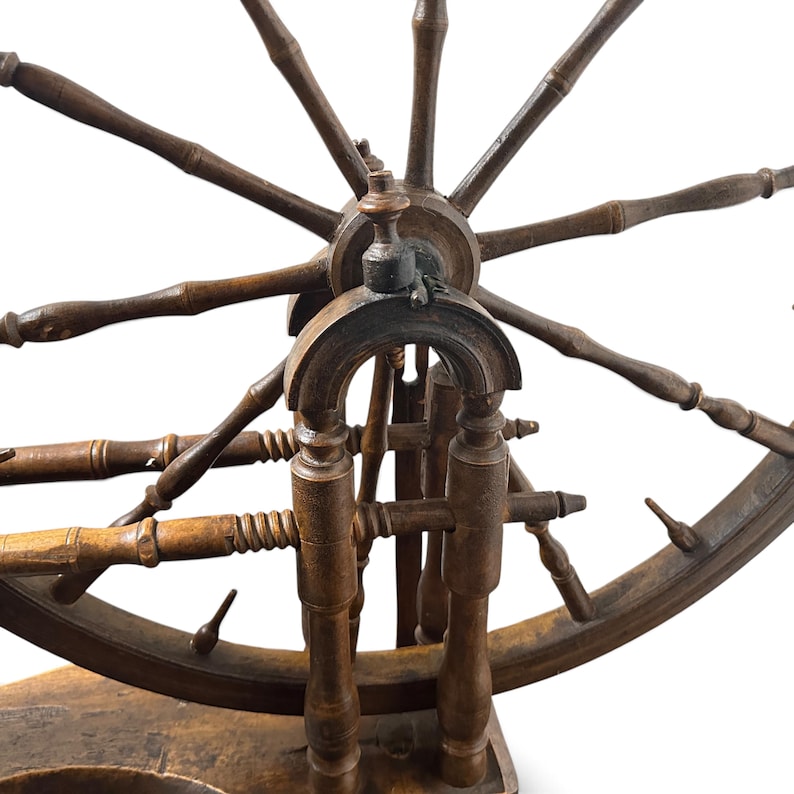 May include: A dark brown wooden spinning wheel featuring a large circular wheel with multiple spokes, a central hub, and a sturdy base. The structure includes turned wooden supports and a horizontal spindle assembly with detailed carved accents.