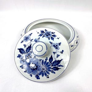 May include: A white ceramic serving dish with a lid, decorated with blue floral patterns. The lid has a central knob and a detailed floral design. The dish's rim is also decorated with blue accents, creating a cohesive and elegant appearance.