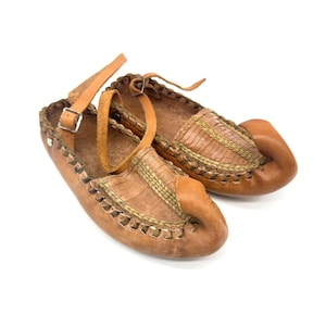 May include: A pair of tan leather shoes featuring a pointed, curled toe design and woven detailing across the top. The shoes include an adjustable ankle strap with a metal buckle and a braided leather trim along the edges of the opening.