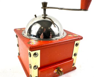 May include: A square wooden coffee grinder painted orange with hand-painted white and yellow daisy motifs. It features a polished silver-tone metal dome, a metal crank handle with a wooden knob, and gold-tone metal corner plates with rivets.