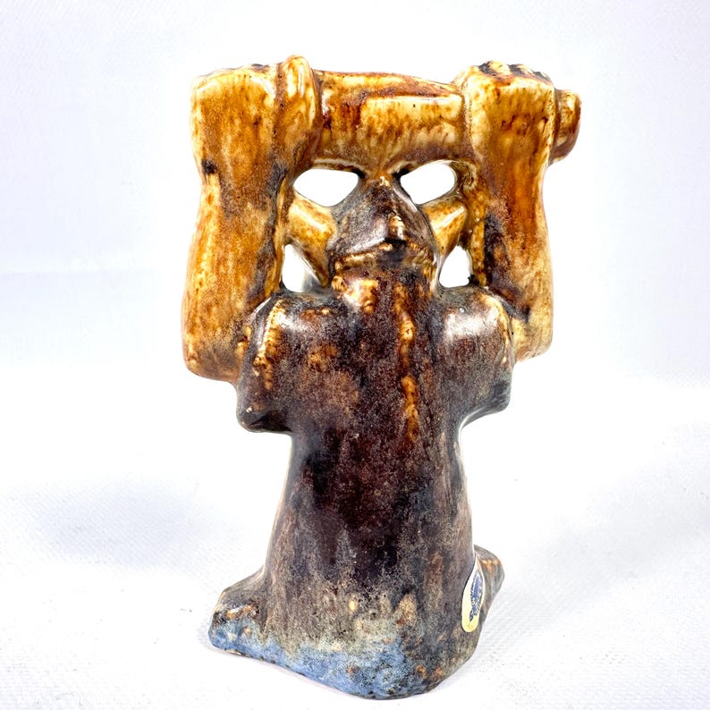 May include: A ceramic figurine with a brown and tan glaze. The figure has a stylized face and is holding a rectangular object above its head. The base of the figurine has a blue hue.