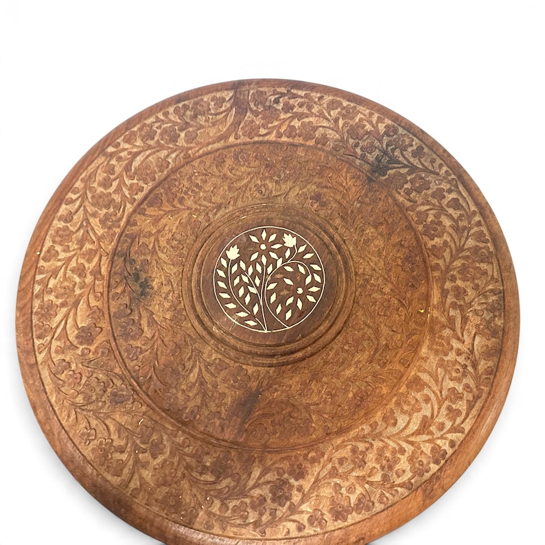 May include: A round wooden tray featuring an intricate carved floral pattern across its surface. The center of the tray displays a circular inlay of light-colored material depicting a stylized plant with leaves and flowers.