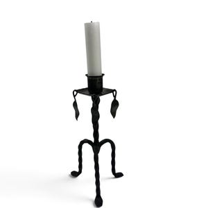 May include: A black metal candle holder with a twisted stem and three curved legs. The top features a square platform with a central cup holding a white pillar candle. Decorative leaf-shaped accents hang from the corners of the square platform.