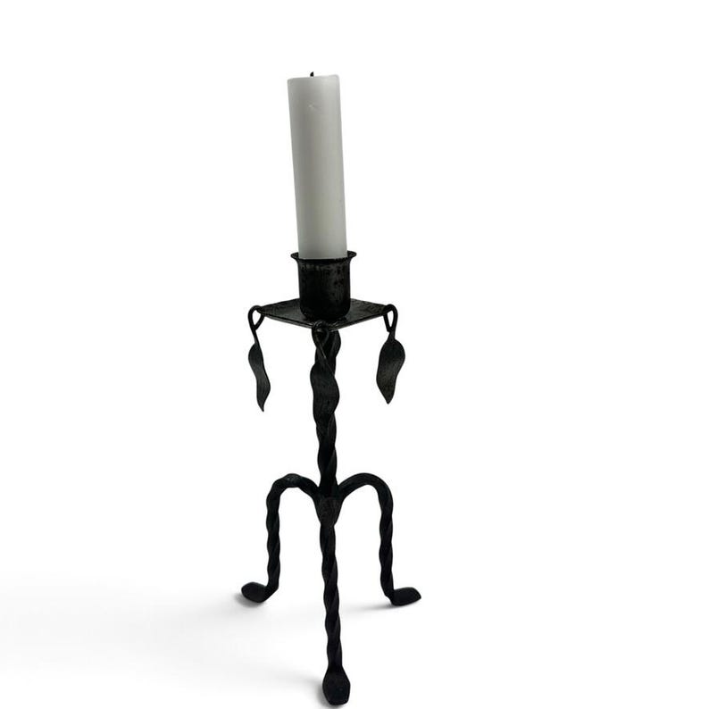 May include: A black metal candle holder with a twisted stem and three curved legs. The top features a square platform with a central cup holding a white pillar candle. Decorative leaf-shaped accents hang from the corners of the square platform.