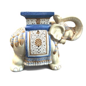 May include: A ceramic elephant garden stool featuring a cream-coloured body with blue and gold accents. The elephant is adorned with an intricate white filigree saddle and a flat blue top surface. It displays detailed tassels and scrollwork.