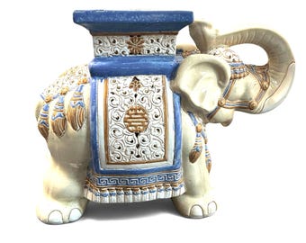 Op de afbeelding: Een keramische olifant-tuinkruk met een crèmekleurig lichaam en blauwe en gouden accenten. De olifant is versierd met een ingewikkeld wit filigraan zadel en een plat blauw bovenoppervlak. Het vertoont gedetailleerde kwastjes en krulwerk.