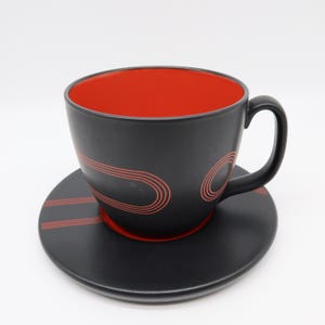 May include: A black teacup and saucer set with a red interior. The cup and saucer have a matte finish and are decorated with red curved lines. The cup has a rounded handle. The saucer is round and flat.
