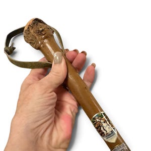 Puede incluir: Un bast&oacute;n de madera marr&oacute;n con un mango natural texturizado y una correa de cuero para la mu&ntilde;eca. El eje est&aacute; adornado con un emblema met&aacute;lico decorativo que representa una cascada y el texto Triberger Wasserf&auml;lle, junto a una segunda etiqueta parcial.