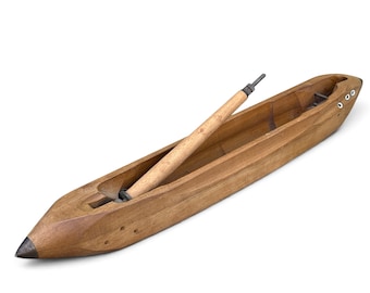 May include: A vintage wooden weaving shuttle with a smooth, polished finish and a pointed metal tip. The elongated, boat-shaped tool features a hollow center containing a removable wooden bobbin, with small metal eyelets visible on the side.