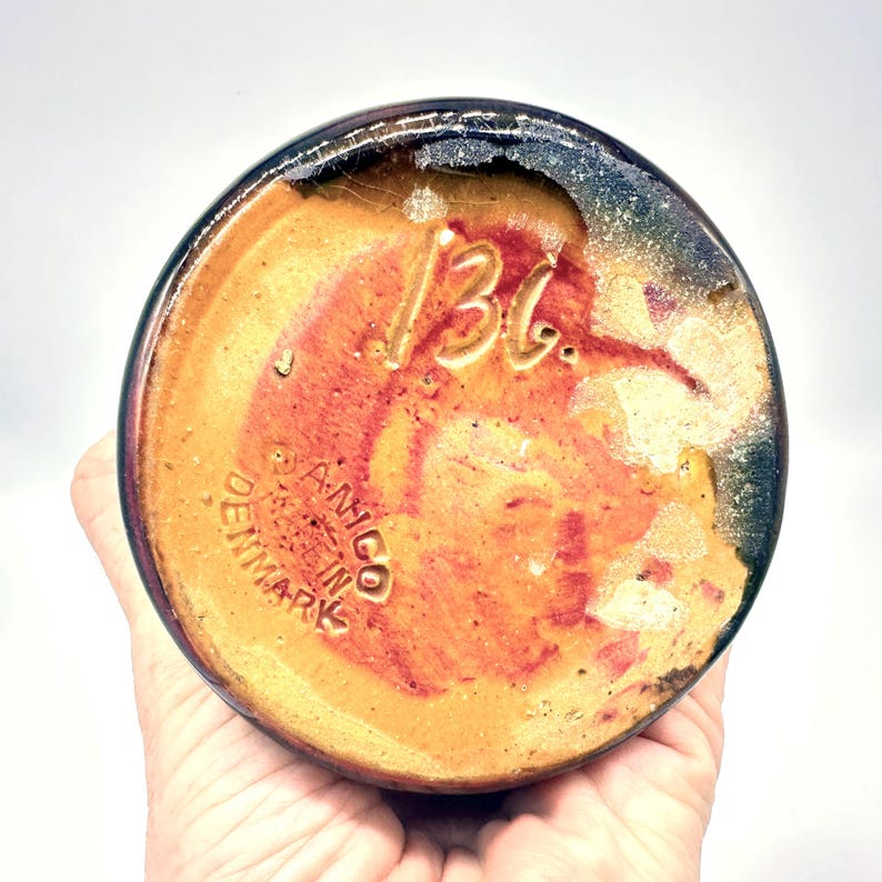 May include: Bottom view of a ceramic vase with a textured, multi-toned surface. The base is primarily orange and brown, with darker blue and black accents around the rim. The number "136" is etched into the base, along with the text "A-NICO DENMARK MADE IN DENMARK".