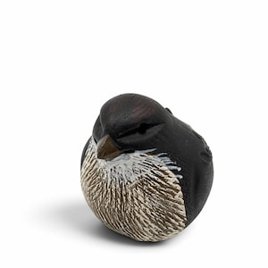 May include: A small, rounded bird figurine with a matte black head and back. The chest area features textured, carved detailing in shades of brown and white. The bird has a small, pointed brown beak and carved eye sockets, presented in a compact, spherical shape.