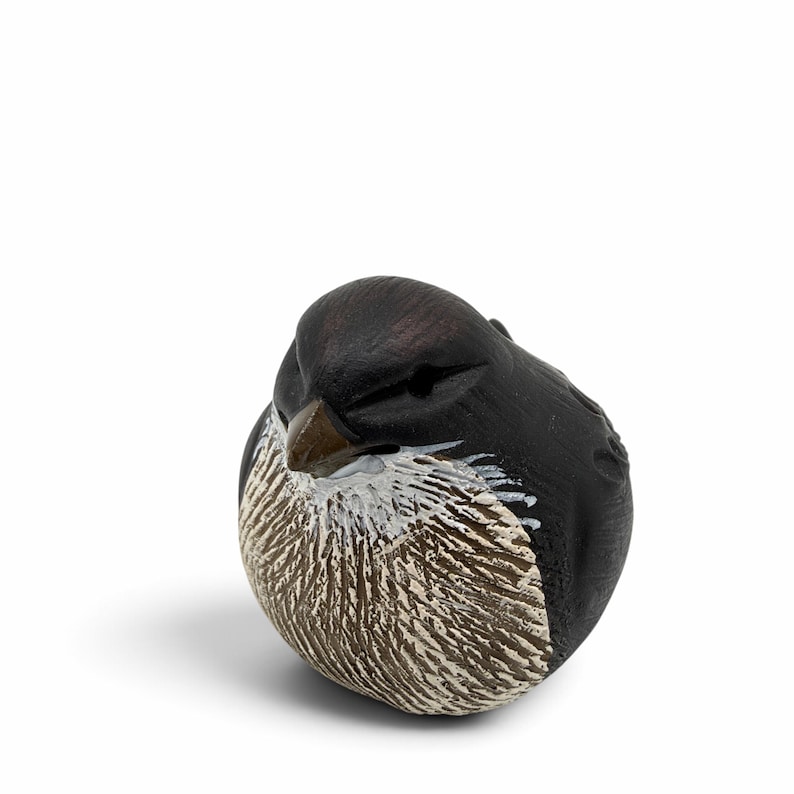 May include: A small, rounded bird figurine with a matte black head and back. The chest area features textured, carved detailing in shades of brown and white. The bird has a small, pointed brown beak and carved eye sockets, presented in a compact, spherical shape.
