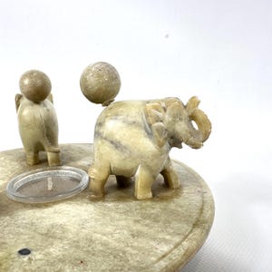 May include: A carved stone elephant figurine with a ball on its back, a small figure, and a clear candle holder. The elephant is light beige with darker veining. The figures are on a round, light beige stone base.