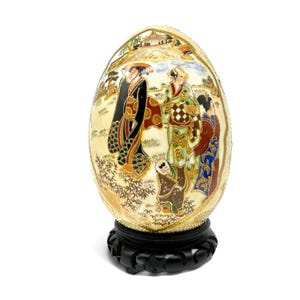 Op de afbeelding: Een eivormig decoratief object met een crèmekleurig oppervlak met een gedetailleerde handgeschilderde scène van figuren in traditionele kleding, inclusief kimono's, tegen een landschap met een gebouw. Het rust op een zwarte houten standaard.