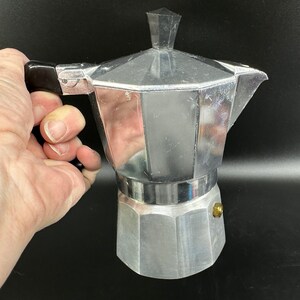 Puede incluir: Una cafetera de aluminio plateado con un mango negro. La cafetera tiene una parte superior redondeada y una base hexagonal.
