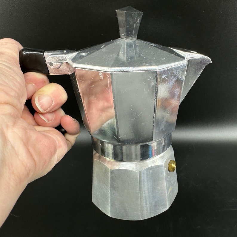 Puede incluir: Una cafetera de aluminio plateado con un mango negro. La cafetera tiene una parte superior redondeada y una base hexagonal.