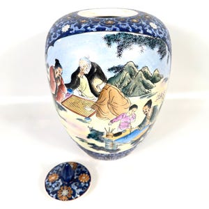 Puede incluir: Un tarro de jengibre de cer&aacute;mica con una escena pintada a mano de figuras jugando a un juego de mesa y practicando caligraf&iacute;a. El tarro tiene un borde y una base con motivos florales en azul y blanco, e incluye una tapa a juego con un remate azul.
