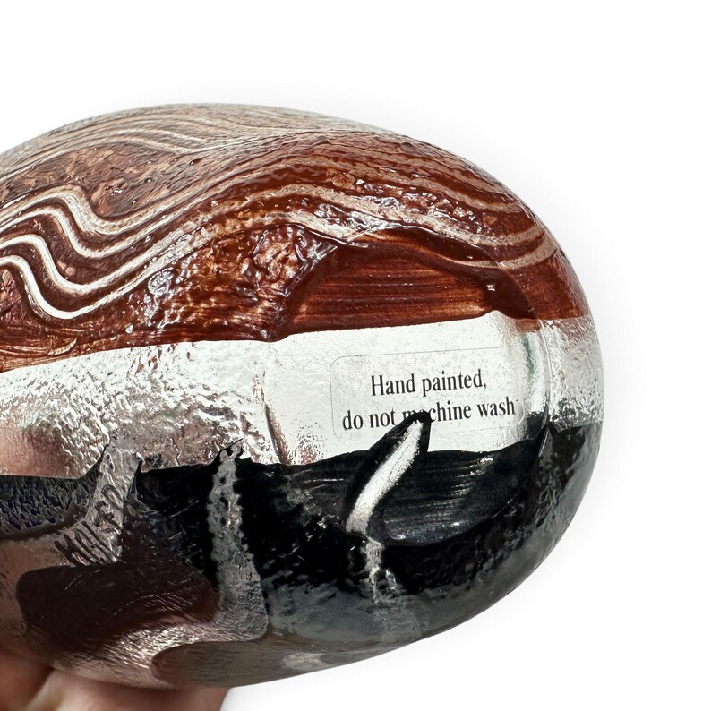 May include: A rounded object featuring a hand-painted design with wavy brown and white lines on the upper portion and solid black on the lower section. A white rectangular label is affixed to the center with the text Hand painted, do not machine wash.