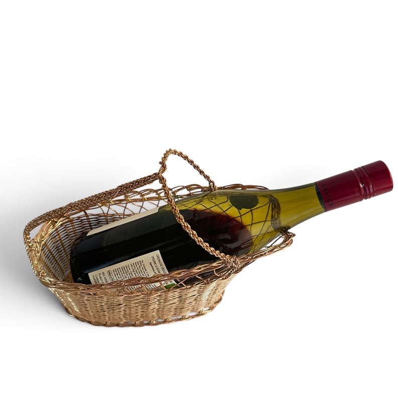 Puede incluir: Una cesta de alambre en tono dorado con base ovalada y un asa arqueada, que contiene una botella de vino de cristal con l&iacute;quido rojo oscuro y un tap&oacute;n de rosca rojo intenso. La botella descansa horizontalmente en la estructura.