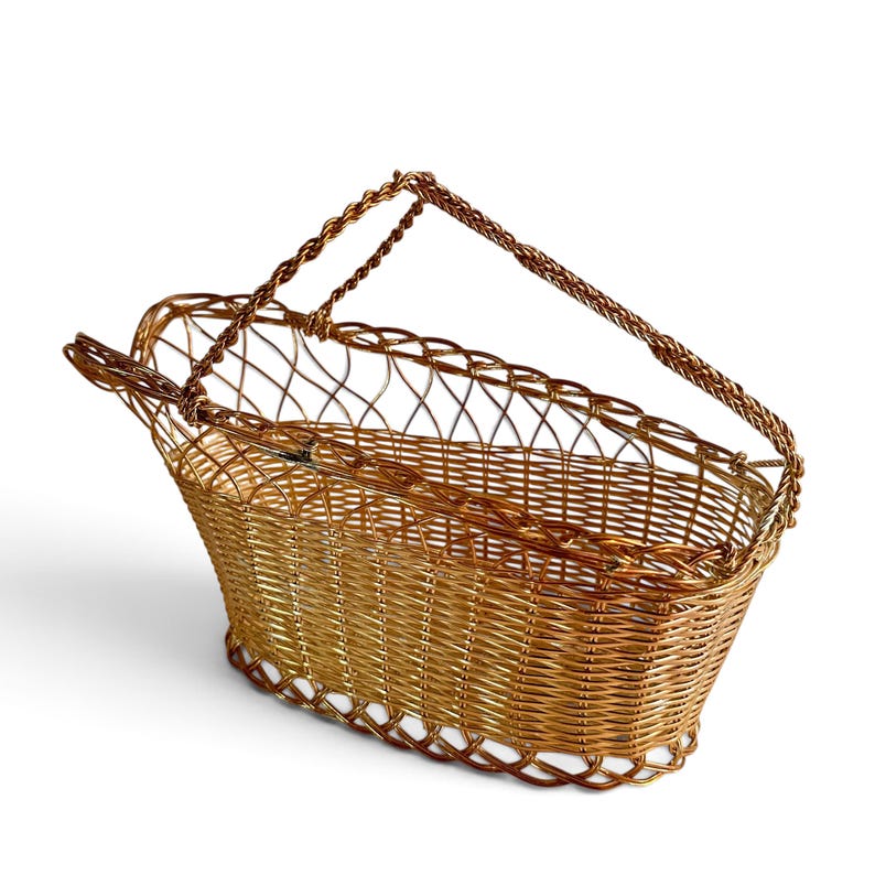 Puede incluir: Una cesta de forma ovalada construida con alambre met&aacute;lico de tono dorado. La cesta presenta una base y laterales tejidos con un patr&oacute;n de celos&iacute;a calado a lo largo del borde superior, complementado por dos asas de alambre trenzado.