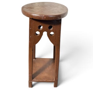 May include: A brown wooden plant stand featuring a circular top, four vertical legs, and a lower square shelf. The upper section of the stand includes decorative cutouts with two circular holes on each side, creating an arched opening.