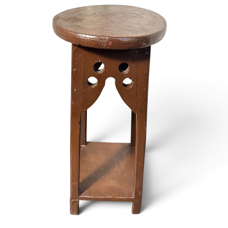 May include: A brown wooden plant stand featuring a circular top, four vertical legs, and a lower square shelf. The upper section of the stand includes decorative cutouts with two circular holes on each side, creating an arched opening.