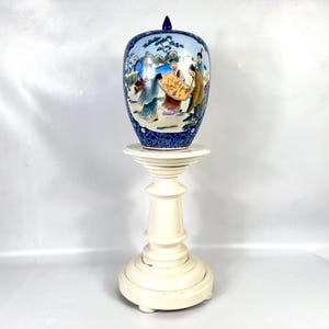 Puede incluir: Un tarro de jengibre de cer&aacute;mica con tapa, que presenta una escena pintada a mano de figuras con t&uacute;nicas tradicionales sobre un fondo azul claro, enmarcado por bordes azul oscuro con motivos florales, sobre un soporte blanco de pedestal.