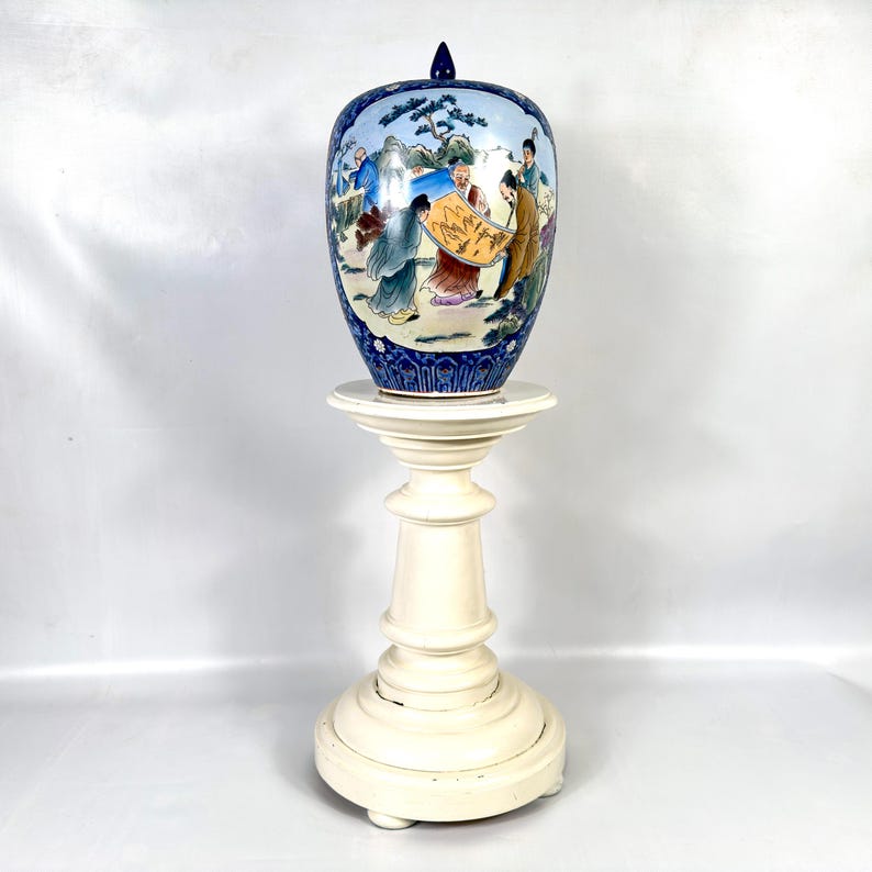 Puede incluir: Un tarro de jengibre de cer&aacute;mica con tapa, que presenta una escena pintada a mano de figuras con t&uacute;nicas tradicionales sobre un fondo azul claro, enmarcado por bordes azul oscuro con motivos florales, sobre un soporte blanco de pedestal.