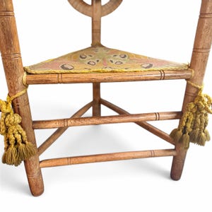 May include: A wooden corner chair featuring a triangular seat upholstered in a yellow and pink moth-patterned fabric. The chair has turned legs and horizontal support rungs, with decorative yellow tassels tied to the front legs.
