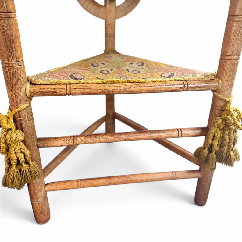May include: A wooden corner chair featuring a triangular seat upholstered in a yellow and pink moth-patterned fabric. The chair has turned legs and horizontal support rungs, with decorative yellow tassels tied to the front legs.