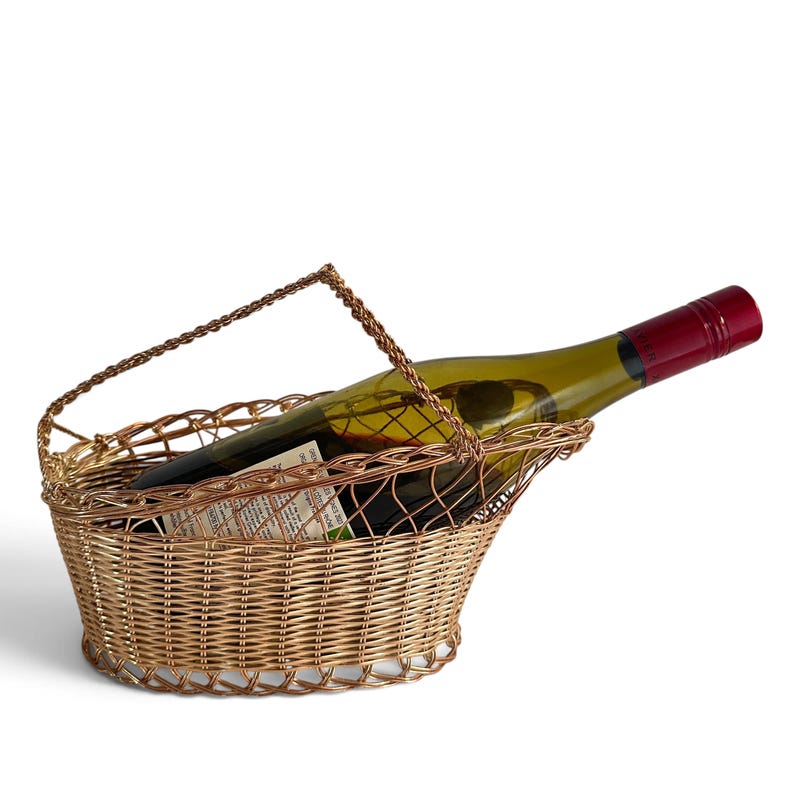 Puede incluir: Un soporte para botella de vino hecho de alambre de color cobre met&aacute;lico, con una base de cesta tejida de forma ovalada y un asa arqueada. Una botella de vino de vidrio con tapa de rosca roja oscura descansa horizontalmente en el marco.