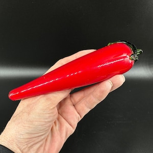 May include: A smooth, elongated red glass sculpture shaped like a chili pepper. The object features a glossy, vibrant red body that tapers to a point, finished with a small, dark green glass stem detail at the wider end, creating a realistic form.