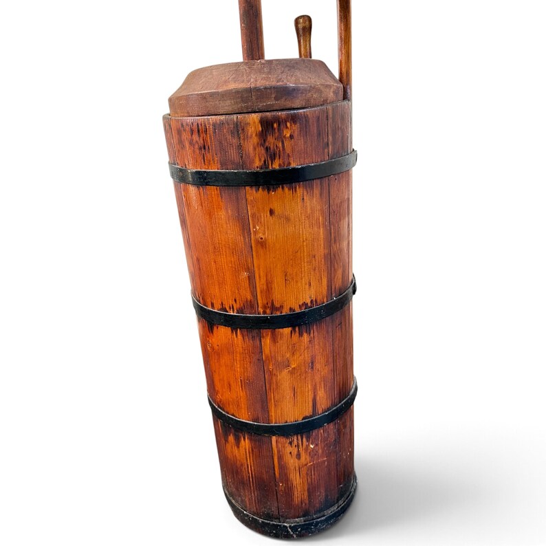 May include: A tall, cylindrical wooden butter churn featuring vertical staves held together by three black metal bands. The churn has a fitted wooden lid with a central opening and a vertical wooden plunger handle extending from the top center.