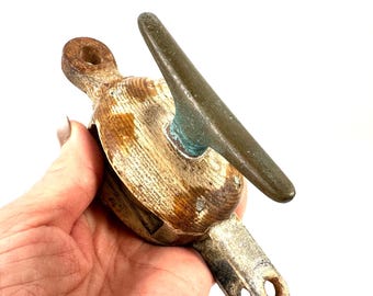 May include: A vintage nautical pulley block featuring a weathered wooden body with visible grain, a dark metallic cleat attached to the side, and a rusted iron attachment point at the top and a dual-holed metal bracket at the bottom end.