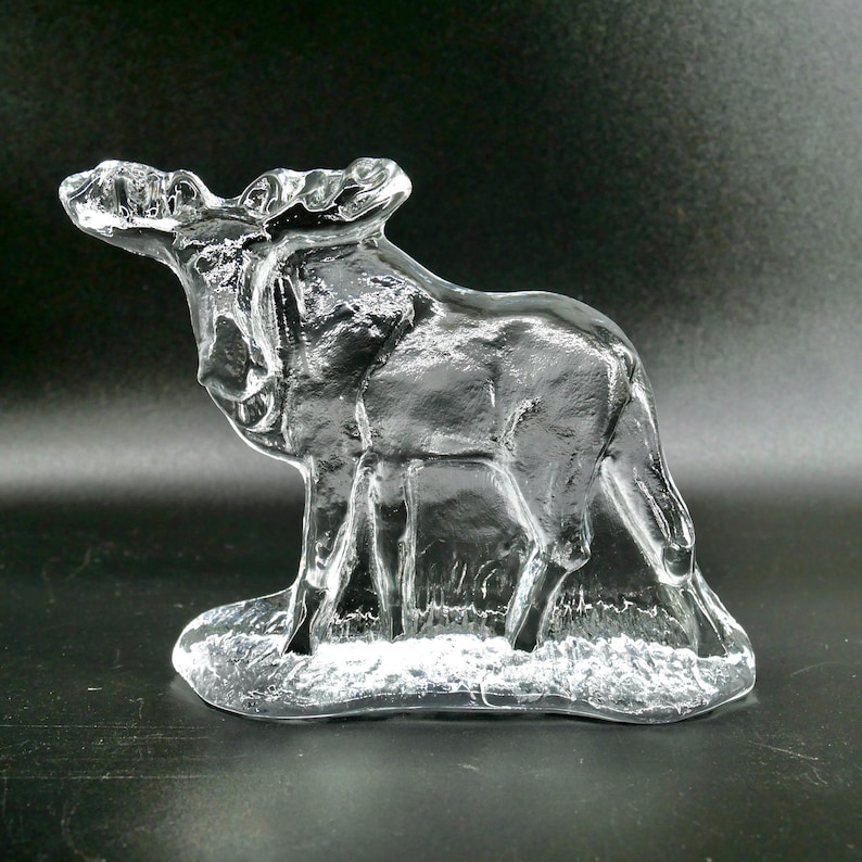 May include: A clear glass moose figurine. The moose is standing on a base, with detailed antlers and body features. The glass is transparent, allowing light to pass through, creating a shimmering effect. The figurine is a decorative item.