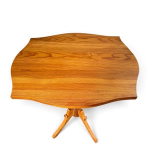 May include: A light brown wooden side table with a smooth, curved, scalloped-edge tabletop featuring a prominent natural wood grain pattern. The table is supported by a central pedestal base that branches into three angled legs.
