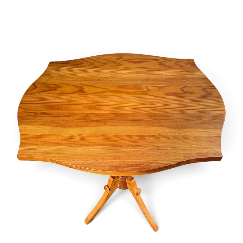 May include: A light brown wooden side table with a smooth, curved, scalloped-edge tabletop featuring a prominent natural wood grain pattern. The table is supported by a central pedestal base that branches into three angled legs.