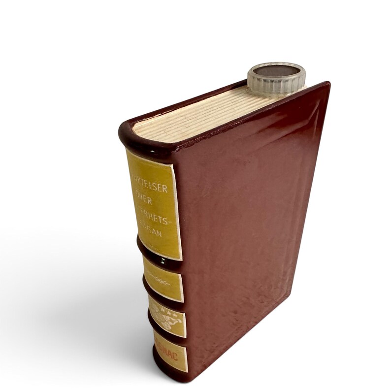 Puede incluir: Una petaca de cer&aacute;mica de color marr&oacute;n oscuro con forma de libro antiguo y un tap&oacute;n de rosca transl&uacute;cido. El lomo presenta etiquetas amarillas con texto blanco, incluyendo las palabras "ERZIELSER", "OVER", "ERRIETS" y "AGAN", y un gr&aacute;fico.