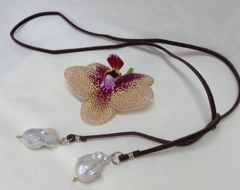 Peut inclure: Un collier en cordon de cuir marron avec deux grosses perles baroques blanches attachées. Le collier est présenté sur une surface blanche avec une orchidée jaune et rouge.