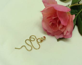 Peut inclure: Un pendentif en fil d'or en forme de ligne ondulée avec un petit charme rond en or au sommet. Le pendentif est posé sur une surface blanche à côté d'une rose rose.
