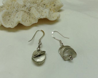 Peut inclure: Boucles d'oreilles argentées avec un design texturé et superposé. Chaque boucle d'oreille présente des éléments en forme de disques superposés, suspendus à un crochet en argent. Une petite perle argentée accentue le design.