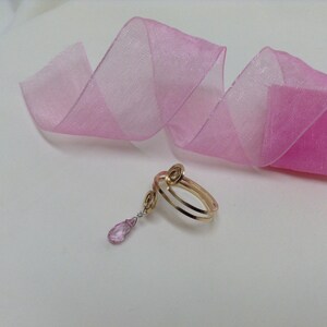 May include: A gold ring with a pink gemstone pendant. The ring is made of two bands of gold that are twisted together. The pendant is a teardrop shape and is attached to the ring by a delicate chain.