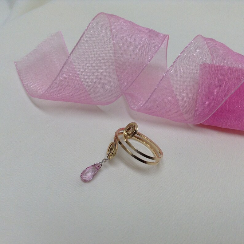 May include: A gold ring with a pink gemstone pendant. The ring is made of two bands of gold that are twisted together. The pendant is a teardrop shape and is attached to the ring by a delicate chain.
