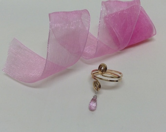 Peut inclure: Une bague en or avec une pierre précieuse rose qui pend. La bague est sur un ruban rose.