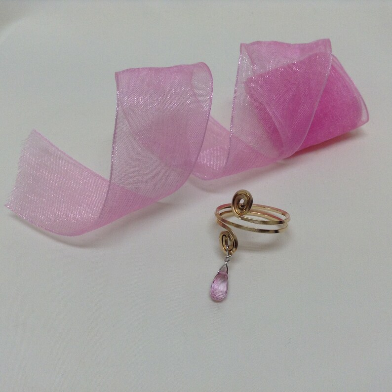 May include: A gold ring with a pink gemstone dangling from it. The ring is on a pink ribbon.