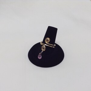 May include: A gold ring with two spirals and a dangling pink gemstone. The ring is displayed on a black ring holder.