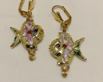 Peut inclure: Une paire de boucles d'oreilles en forme de poisson dorées avec des détails en émail rose, blanc et vert. Les poissons ont un œil rouge et noir et une perle de cristal transparente qui pend.