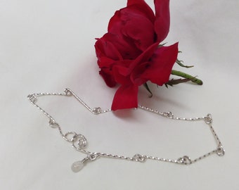 Peut inclure: Un bracelet de cheville en argent avec un petit charme en forme de larme transparente. Le bracelet de cheville est posé sur une surface blanche à côté d'une rose rouge.