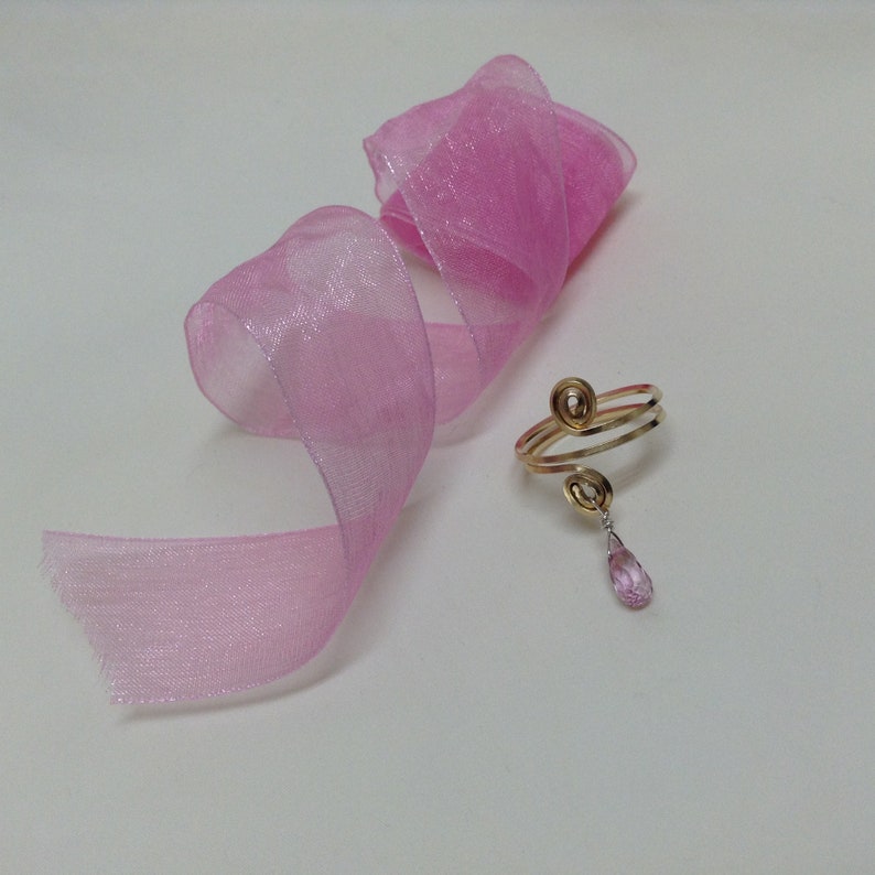 May include: A gold ring with a pink stone dangling from it. The ring is made of two intertwined bands. The ring is on a pink ribbon.