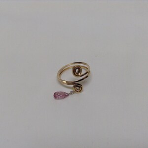 May include: A gold ring with a pink crystal pendant. The ring is made of two intertwined bands of metal, and the pendant is a teardrop-shaped crystal.